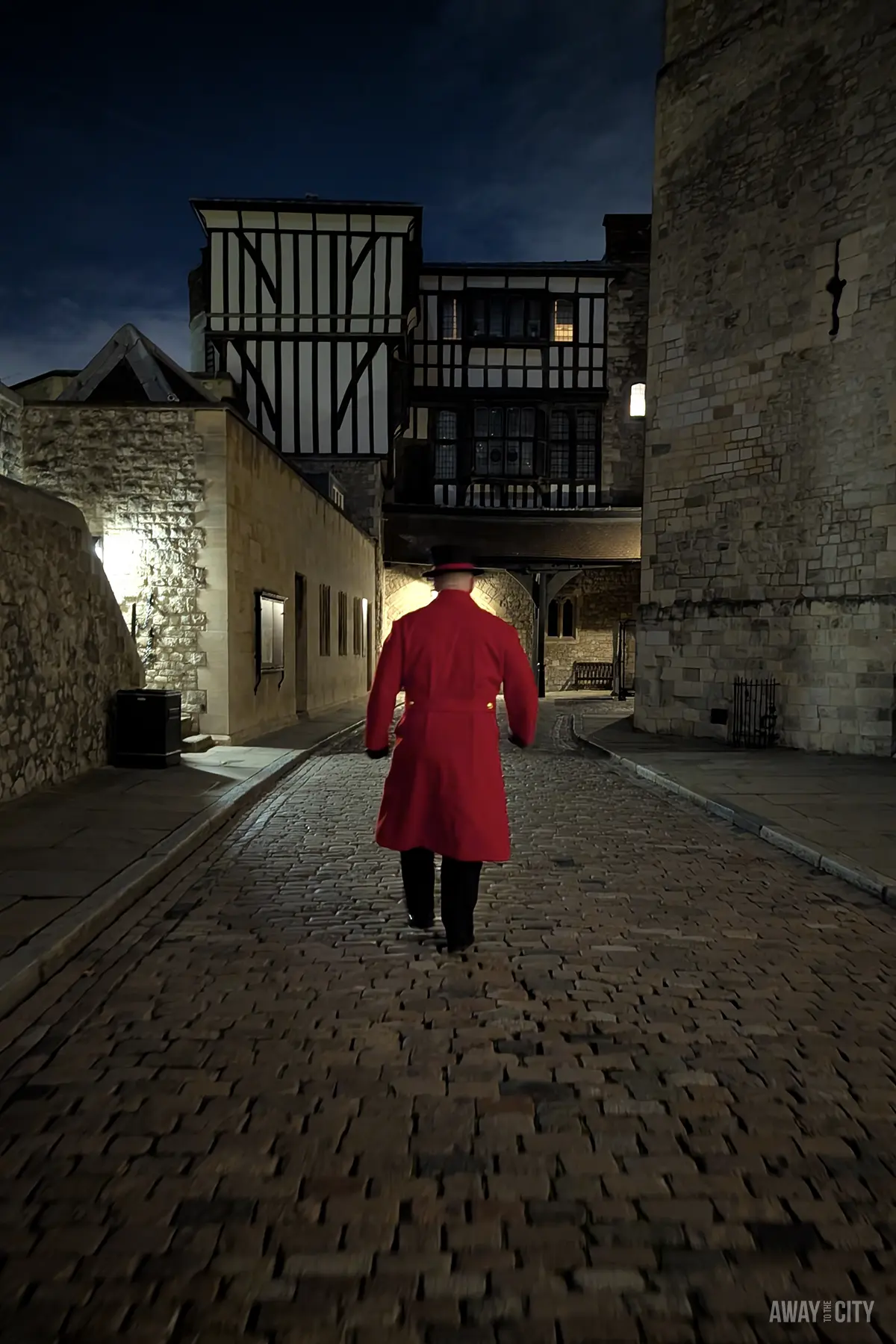 A Yeoman Warder in a red coat is seen from behind, walking up a dark cobblestone street within the Tower of London.