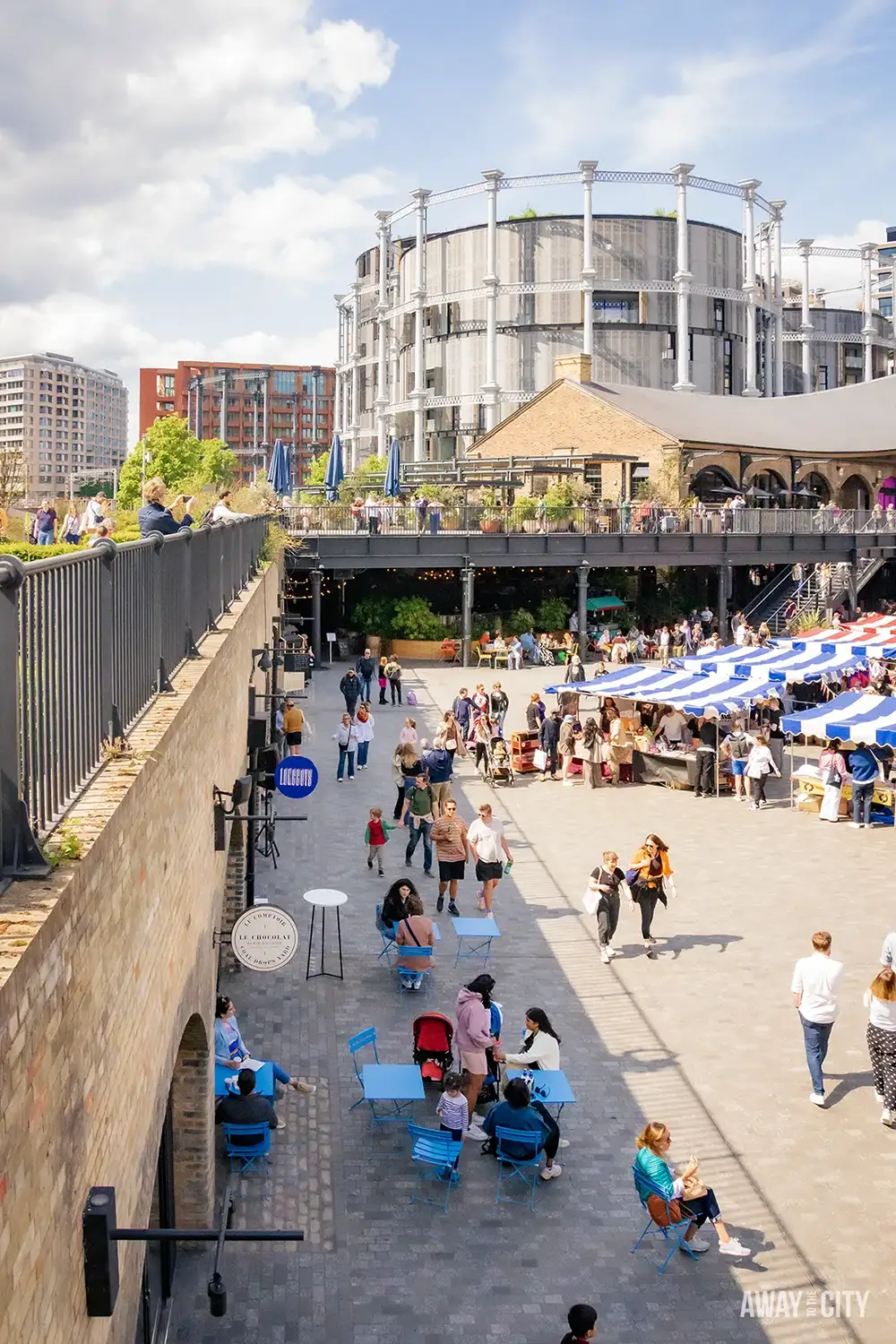 A bustling urban courtyard, Coal Drops Yard, is filled with people, outdoor seating, and shops, with a large, modern circular structure in the background.