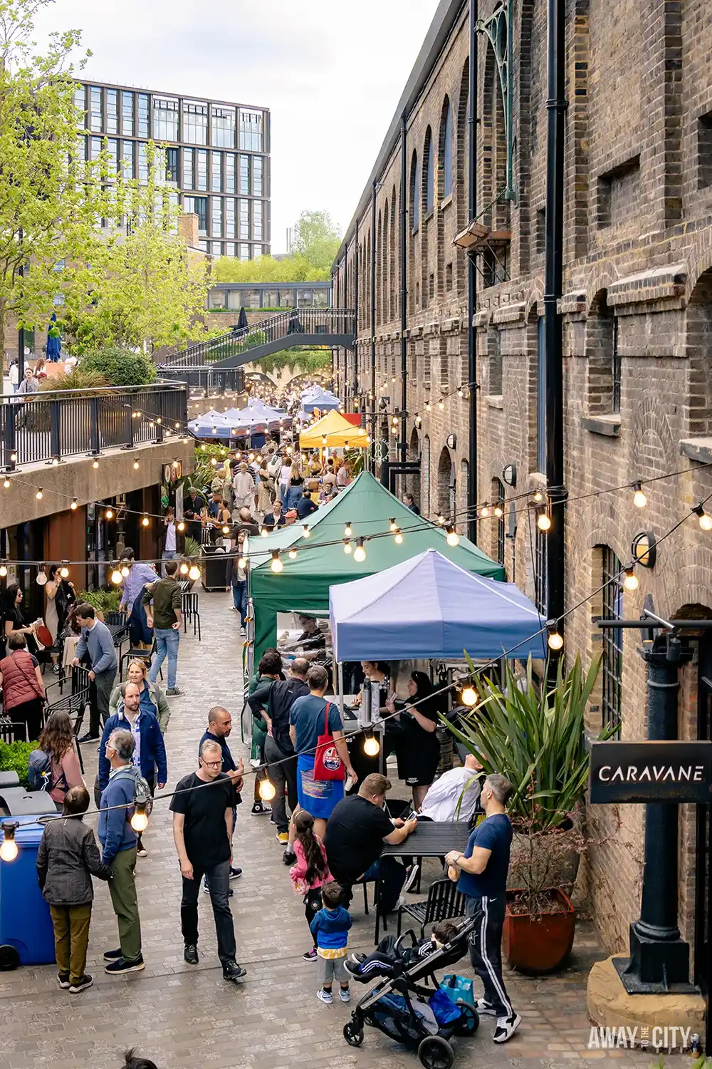 A bustling market street in London's Coal Drops Yard features vendors with striped canopies, string lights, and a crowd of people walking and dining at outdoor tables.