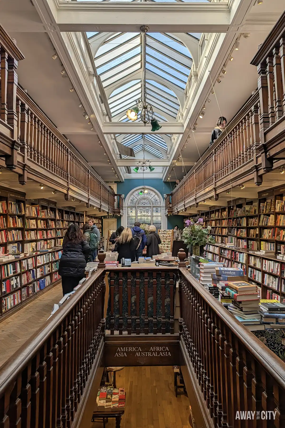 he interior of Daunt Books in London features long wooden galleries with railings, a glass ceiling, and bookshelves lining the walls.