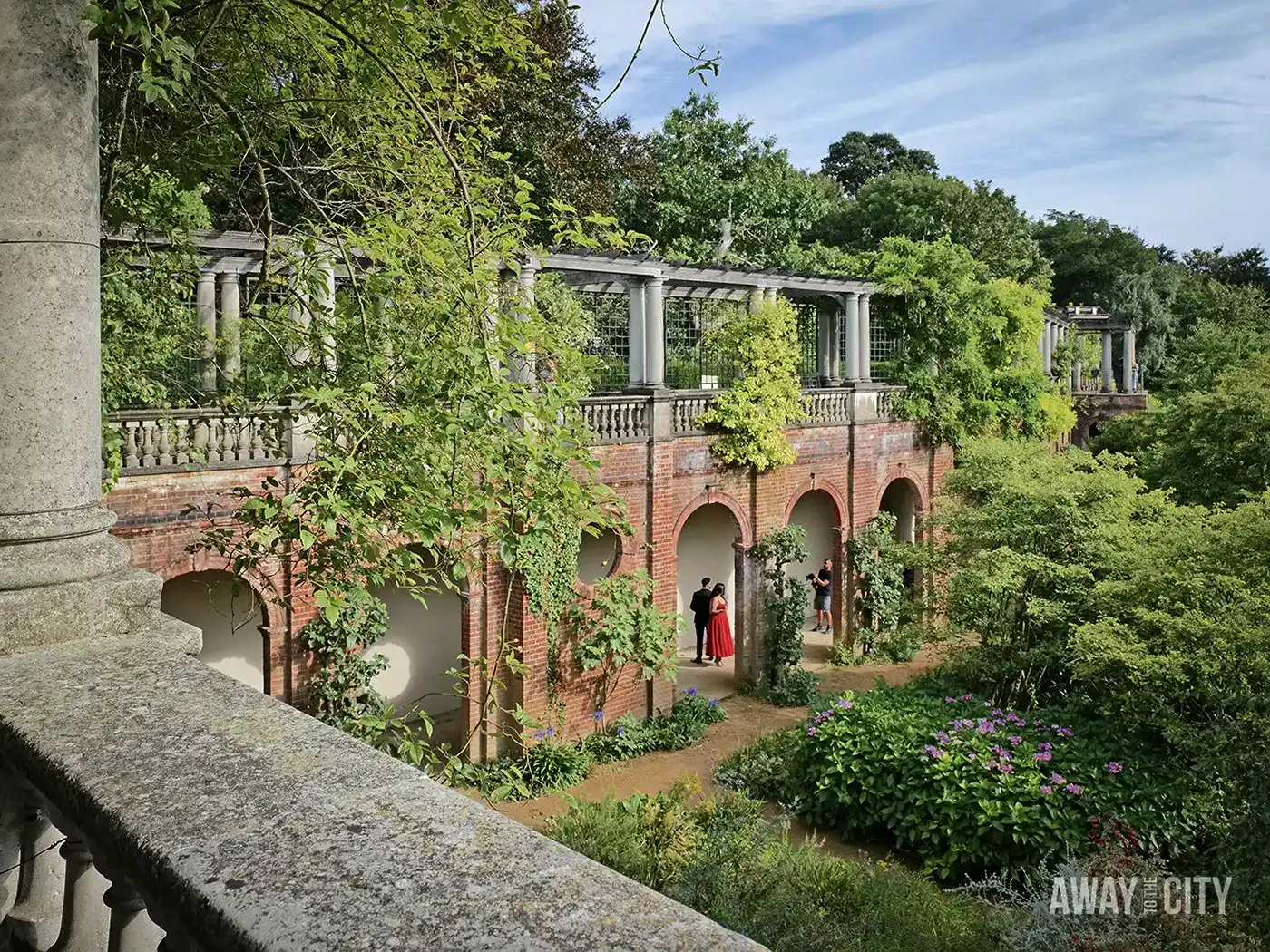 A garden pergola at Hampstead Hill, London, features a long, brick colonnade overgrown with green ivy and foliage, with people visible in the distance.