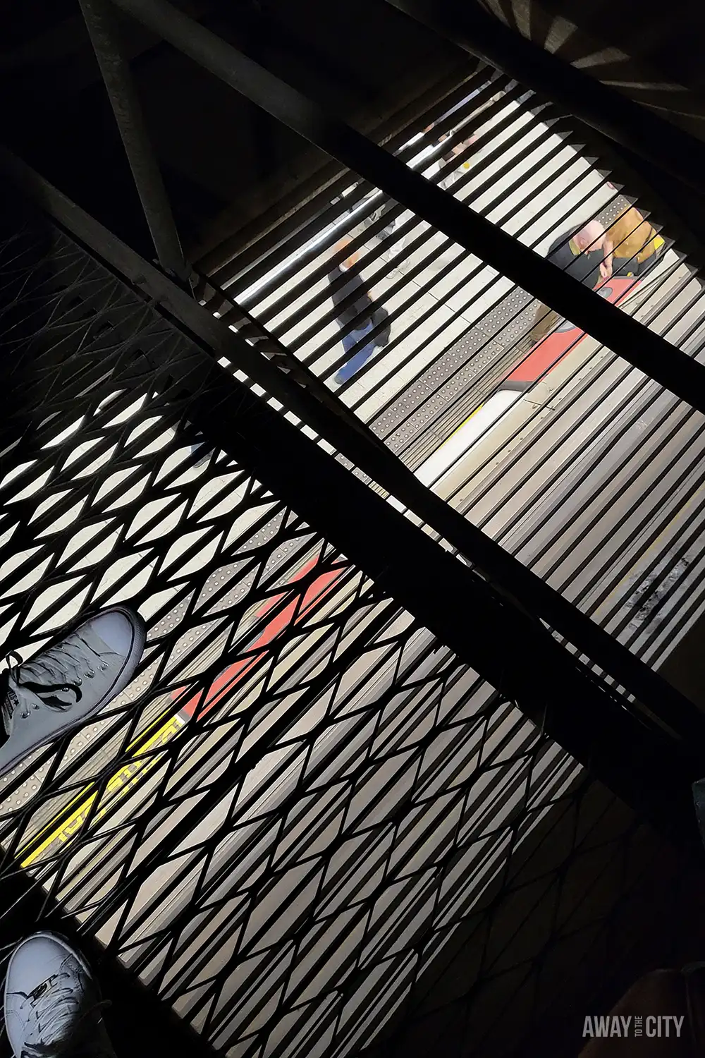 A low-angle view shows two people's feet on a grated metal platform, looking down at a London Underground platform with a train pulling in.