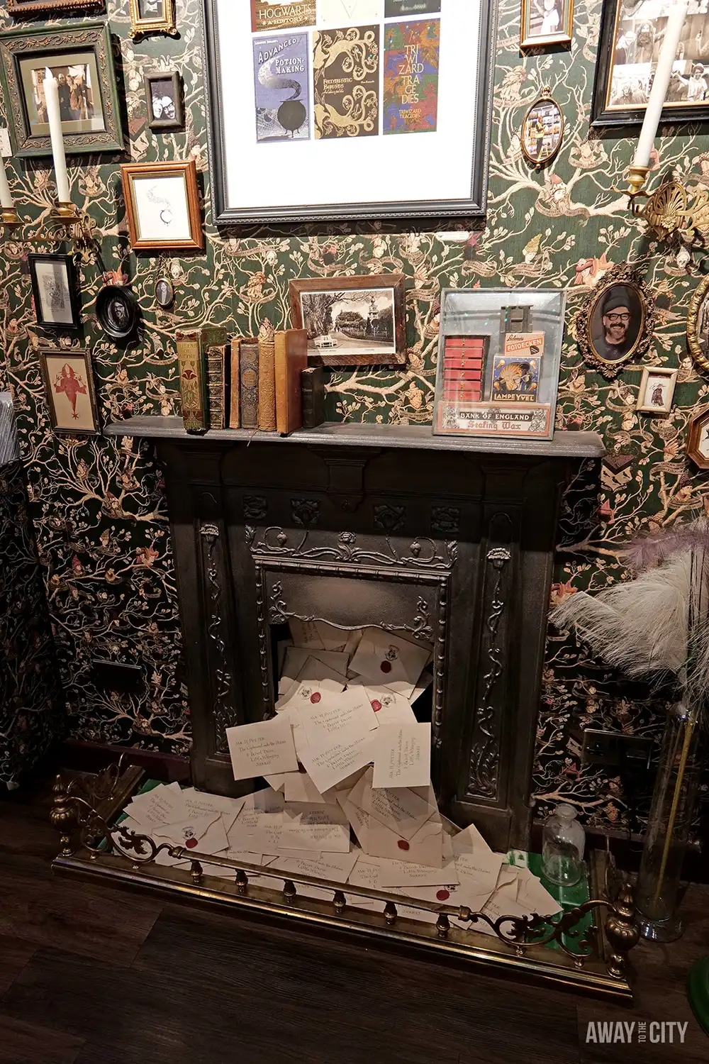 An ornate fireplace is overflowing with a pile of letters, and the mantelpiece above is decorated with books and framed art, set against dark, patterned wallpaper.