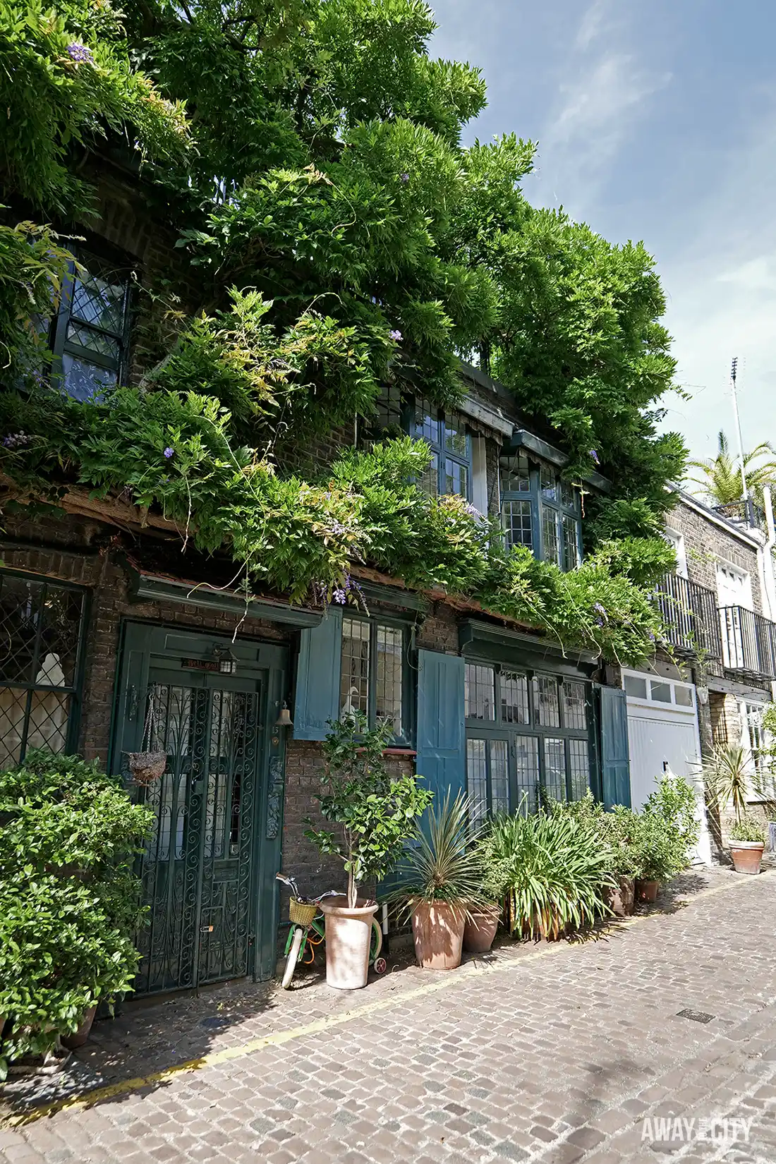A tree-lined cobbled street in London features charming brick houses and abundant green foliage under a bright sky.