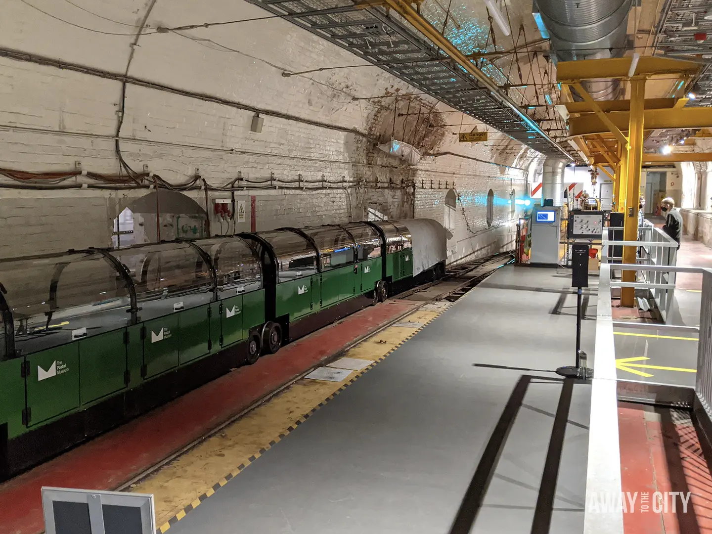 A train from the Postal Museum sits on a track in a well-lit tunnel, with a yellow line on the floor and a tour group visible in the background.