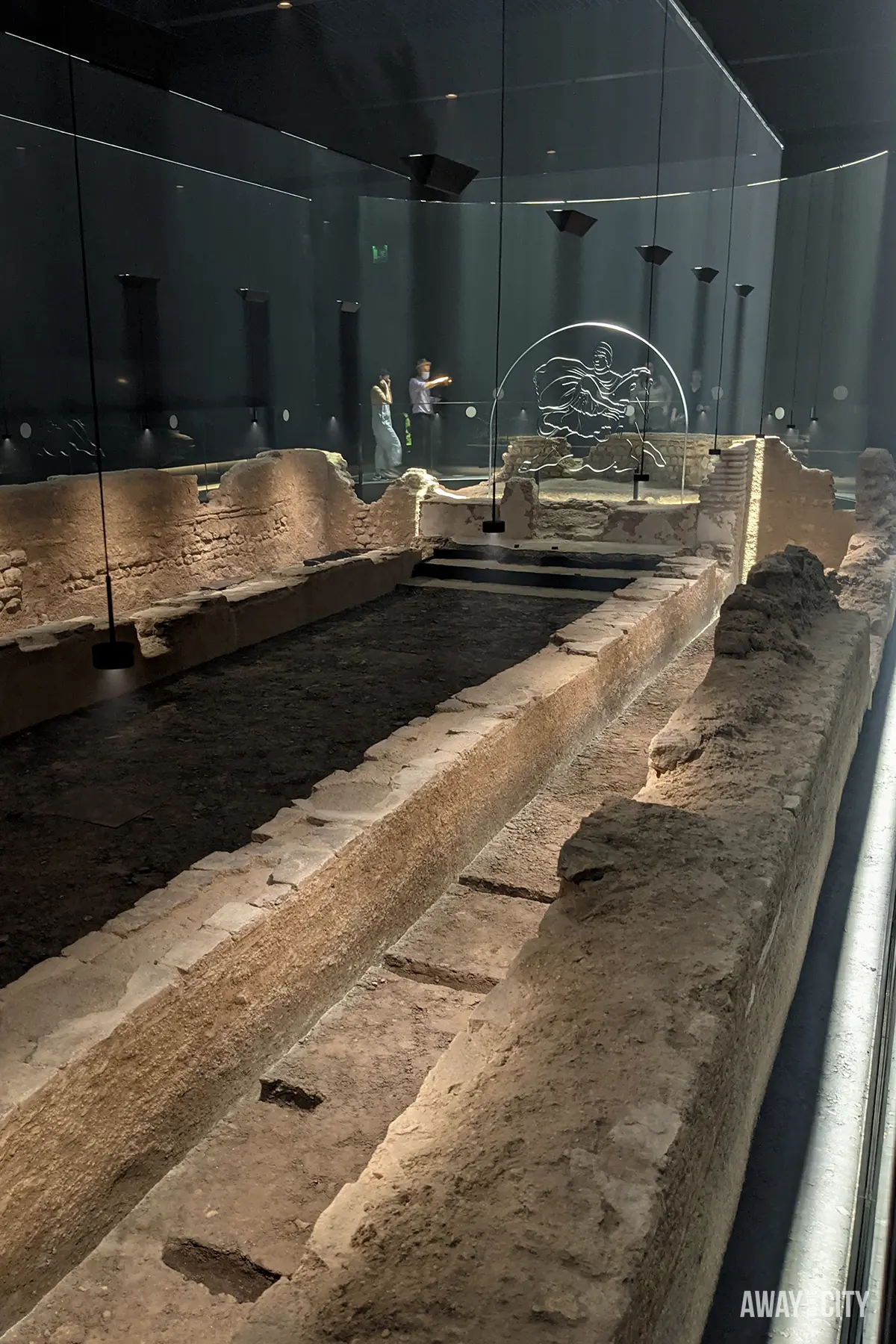 The interior of a museum features a dark room with the excavated remains of a Roman temple, illuminated by spotlights and a light installation outlining a bull sculpture.