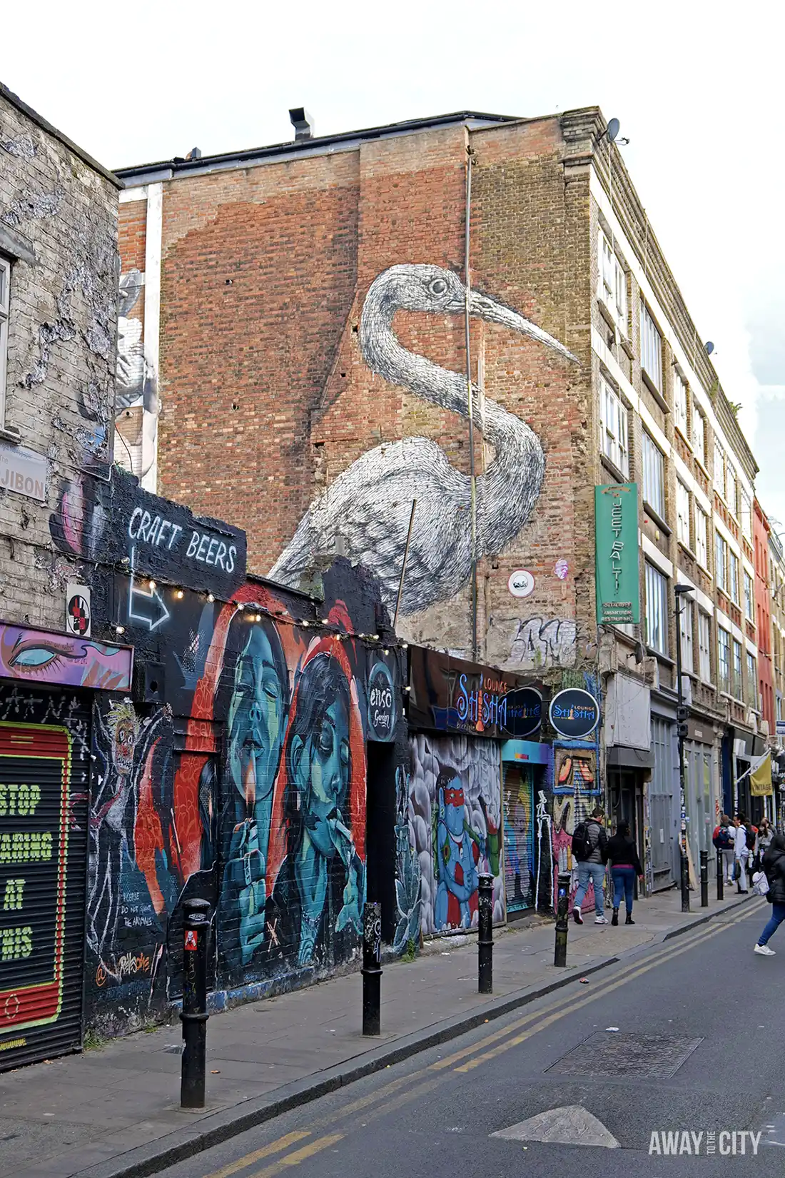 A street in London is lined with multi-colored buildings, with a large white crane graffiti on a brick wall.