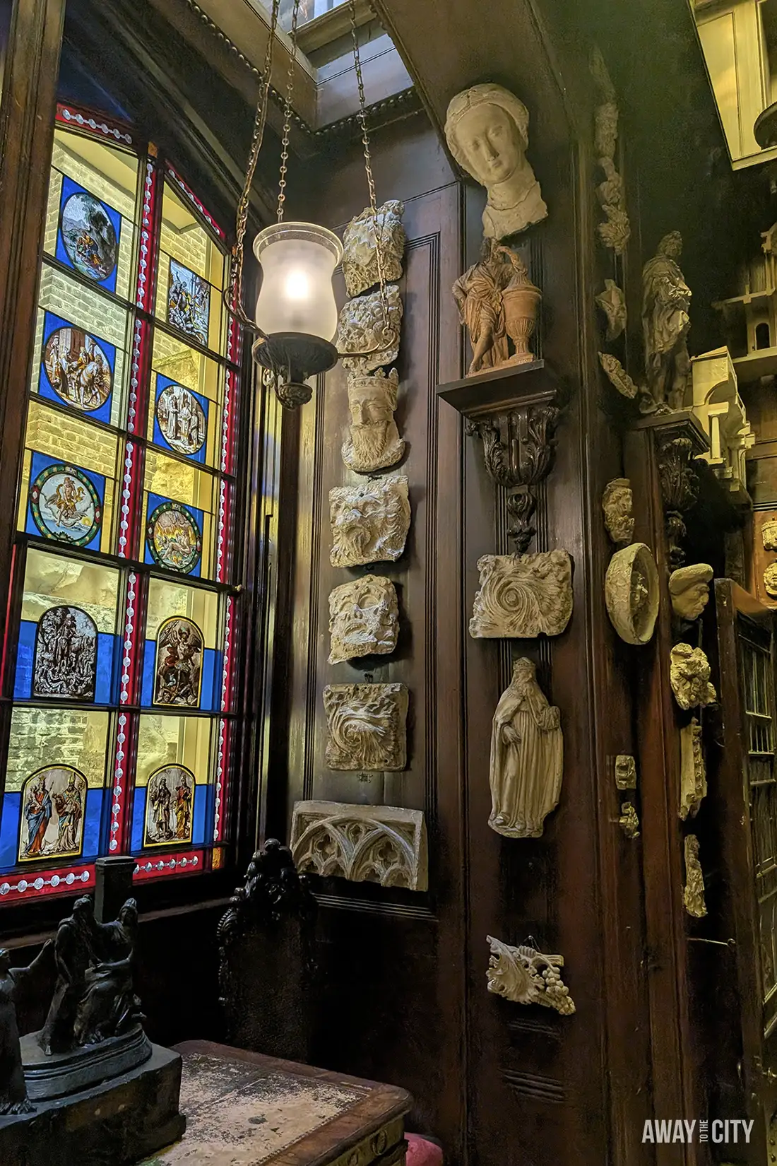 A dark wooden wall in the Sir John Soane's Museum is covered with an eclectic display of classical busts, decorative stone fragments, and sculptures, next to a large stained-glass window.