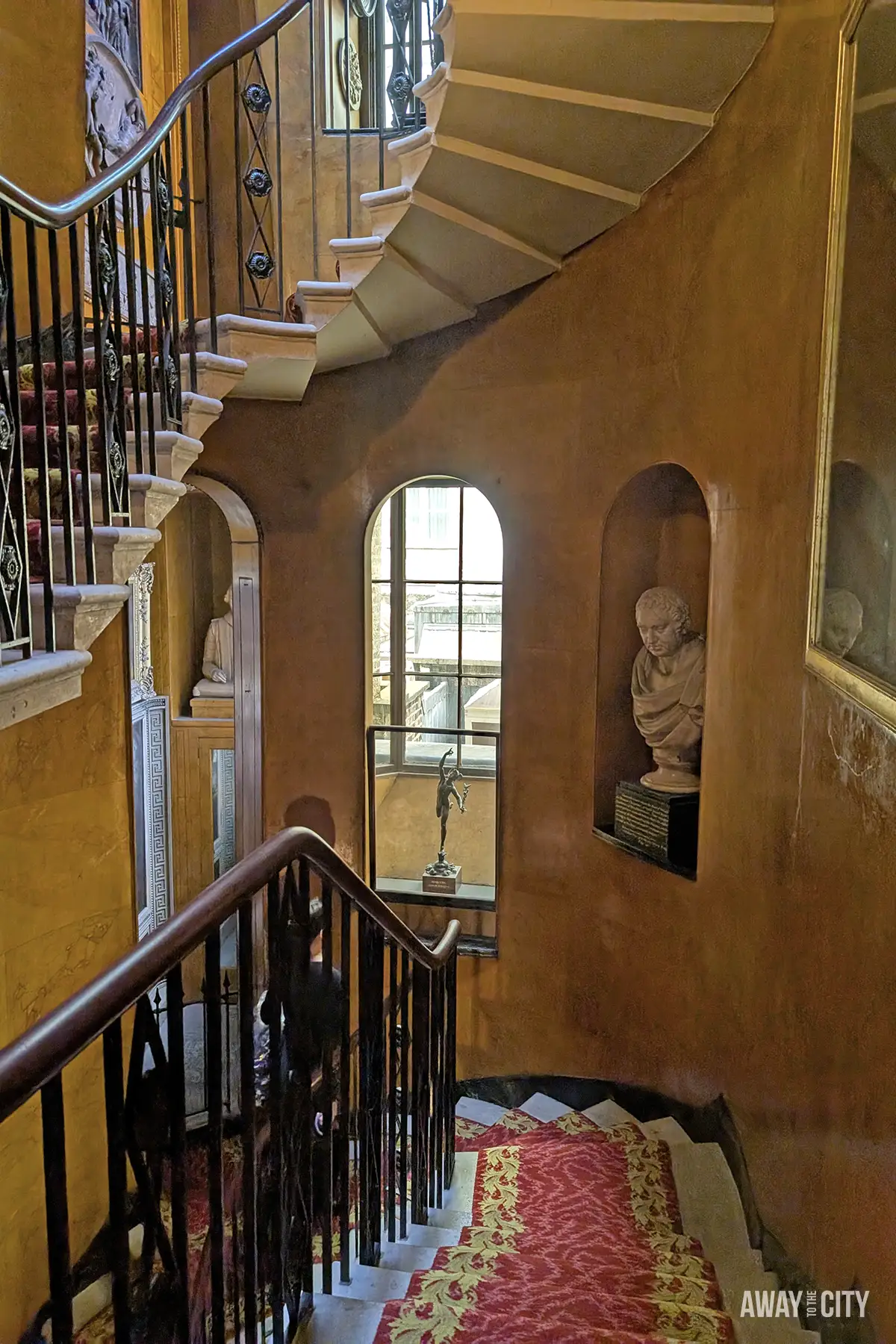 A winding staircase with dark wooden banisters and a red patterned carpet ascends within a dimly lit, orange-walled room, featuring a window and a white bust in a niche.