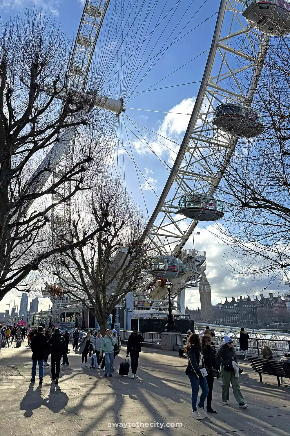 The London Eye stands tall, with its pods visible, and people walk along the riverside path below.