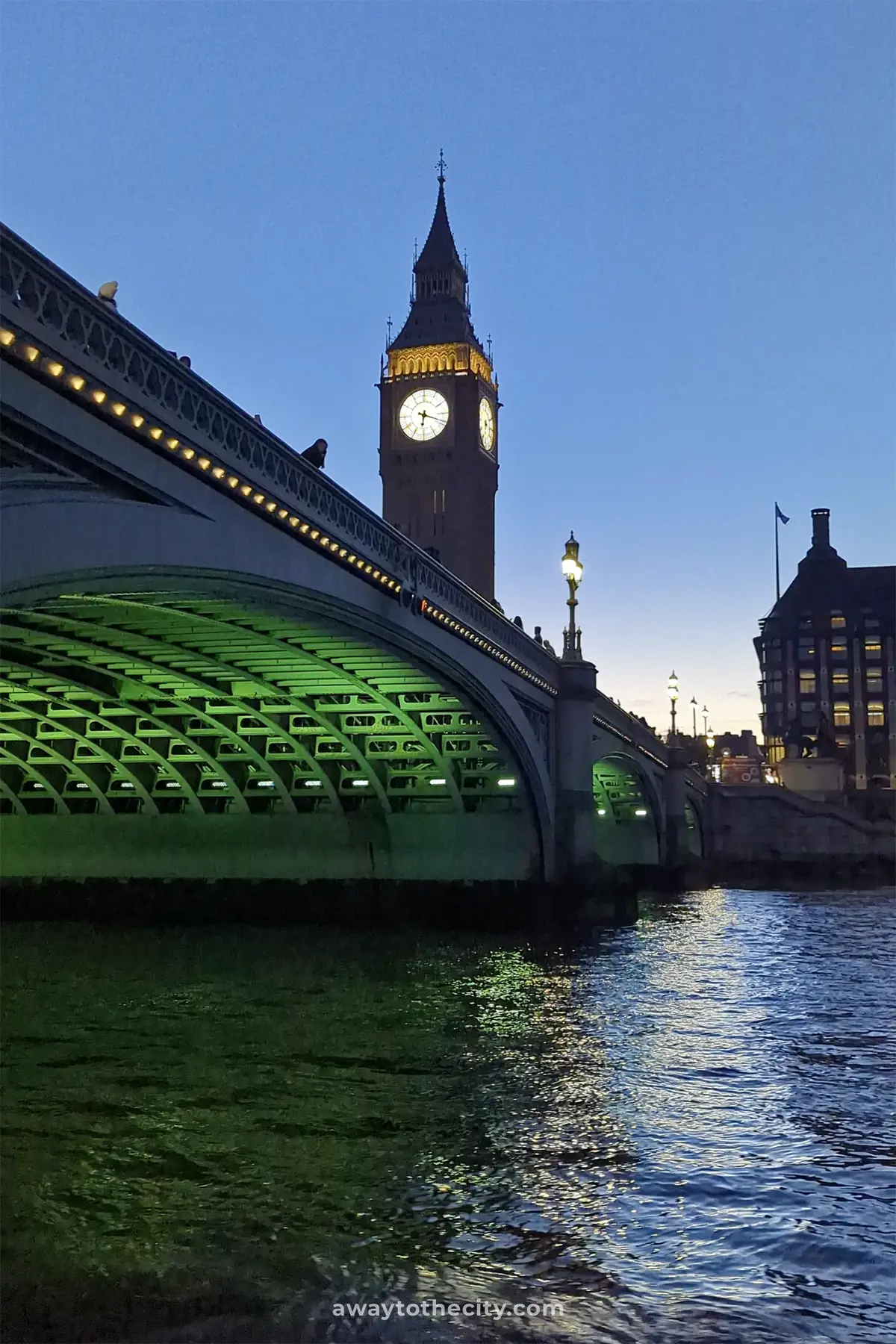 Big Ben is lit up at dusk, seen from the River Thames with a bridge in the foreground.