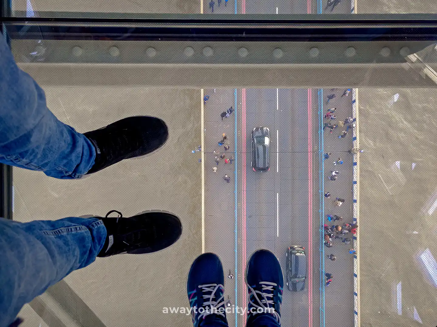 Two pairs of feet are seen on the glass floor of Tower Bridge, looking down at traffic and people below.