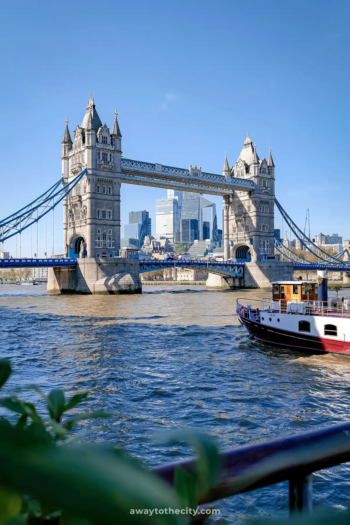 Tower Bridge spans the River Thames on a clear day, with a boat in the foreground and city buildings behind.