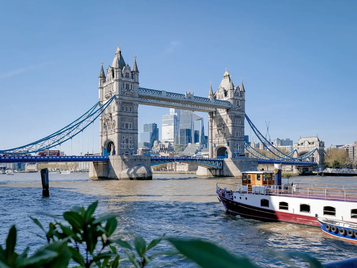 Tower Bridge spans the River Thames on a clear day, with a boat in the foreground and city buildings behind.