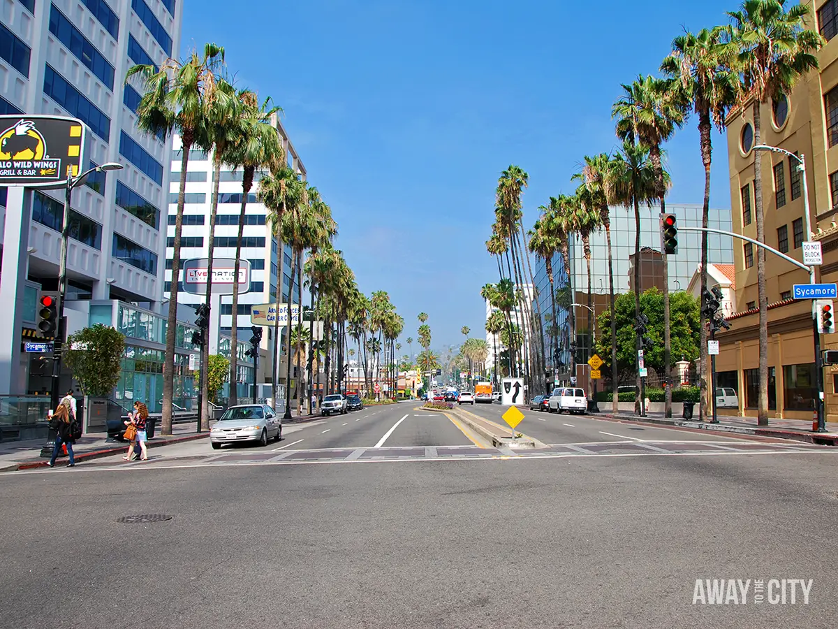 Wide Los Angeles boulevard lined with tall palm trees, modern buildings and light traffic, with visible signs for Buffalo Wild Wings and Sycamore.