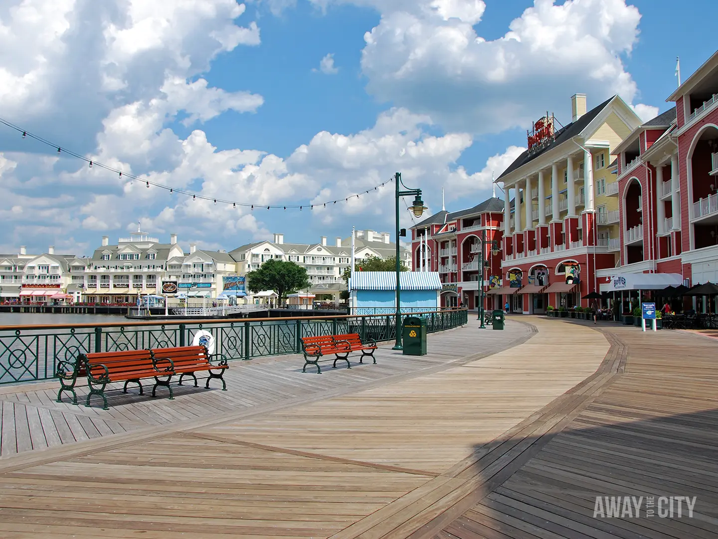 Wooden boardwalk beside calm water with benches, string lights and pastel waterfront buildings under bright blue sky and scattered clouds.
