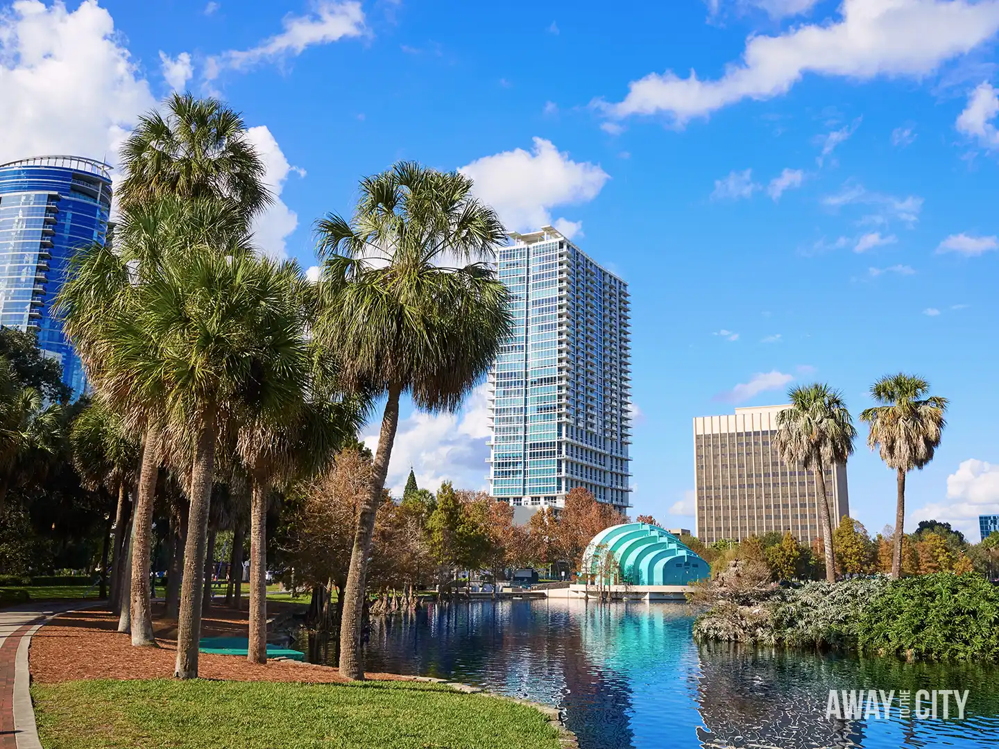 Palm-lined lakeside park with reflective water, curved amphitheatre stage and modern high-rise buildings rising behind green lawns.