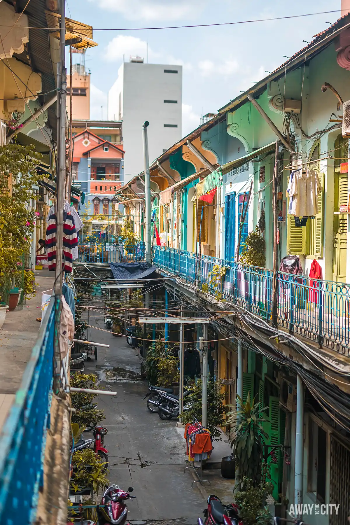 Narrow residential corridor with colourful balconies, hanging laundry, potted plants and overhead cables, looking down onto parked motorbikes below.