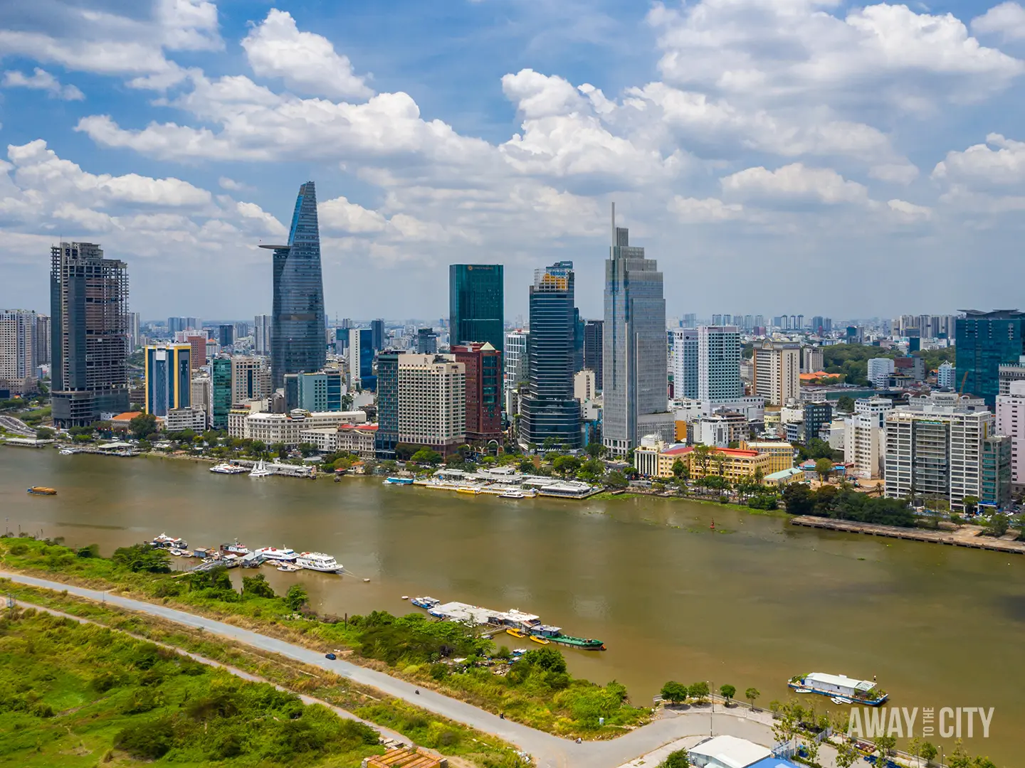 Ho Chi Minh City skyline along the Saigon River, featuring Bitexco Financial Tower, modern high-rises, boats and green riverbanks under cloudy sky.